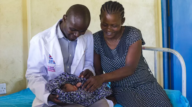 A healthcare provider and woman smile down at a baby during a health visit.